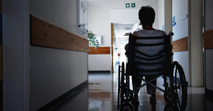 A nursing home resident in a wheelchair sits in an empty hallway.