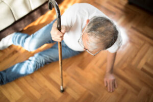 An older man in a nursing home has fallen to the floor, holding himself up with a cane.