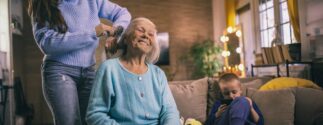 An older woman gets her hair braded by a younger woman with holiday decor in the background.