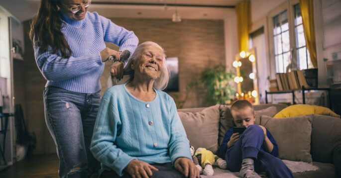 An older woman gets her hair braded by a younger woman with holiday decor in the background.