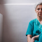 A woman in green scrubs sits against the wall, appearing to be exhausted.