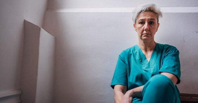 A woman in green scrubs sits against the wall, appearing to be exhausted.