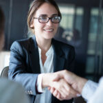 A professional woman with glasses shakes a man's hand across from her, while another woman sits next to him.