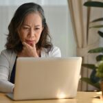 A woman sitting at a table, looking thoughtfully at her laptop screen.